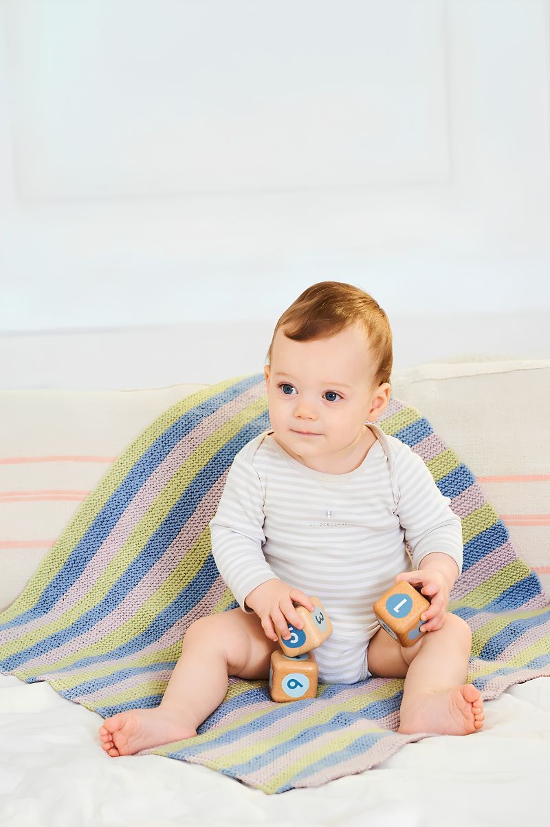 Baby sitting on a striped double knit blanket holding wooden toys against a white background
