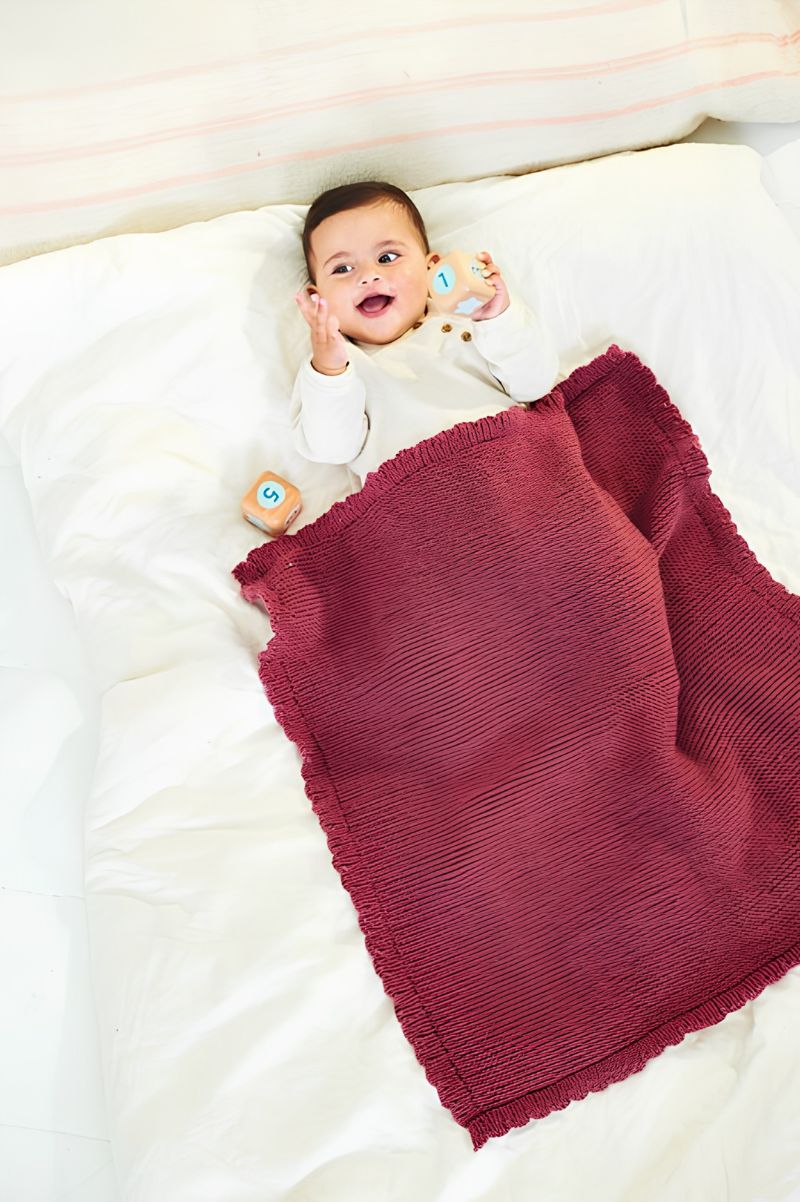 Baby lying on a white blanket with a red knitted blanket draped over them, wearing a white outfit.