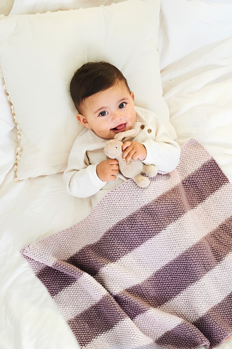 Baby lying on a striped blanket holding a toy, surrounded by soft bedding