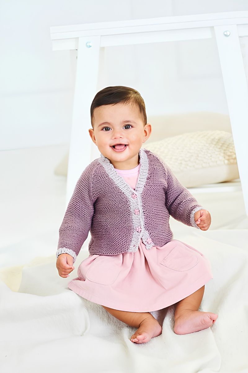 Baby wearing a pink knitted cardigan and skirt sitting on a white blanket.