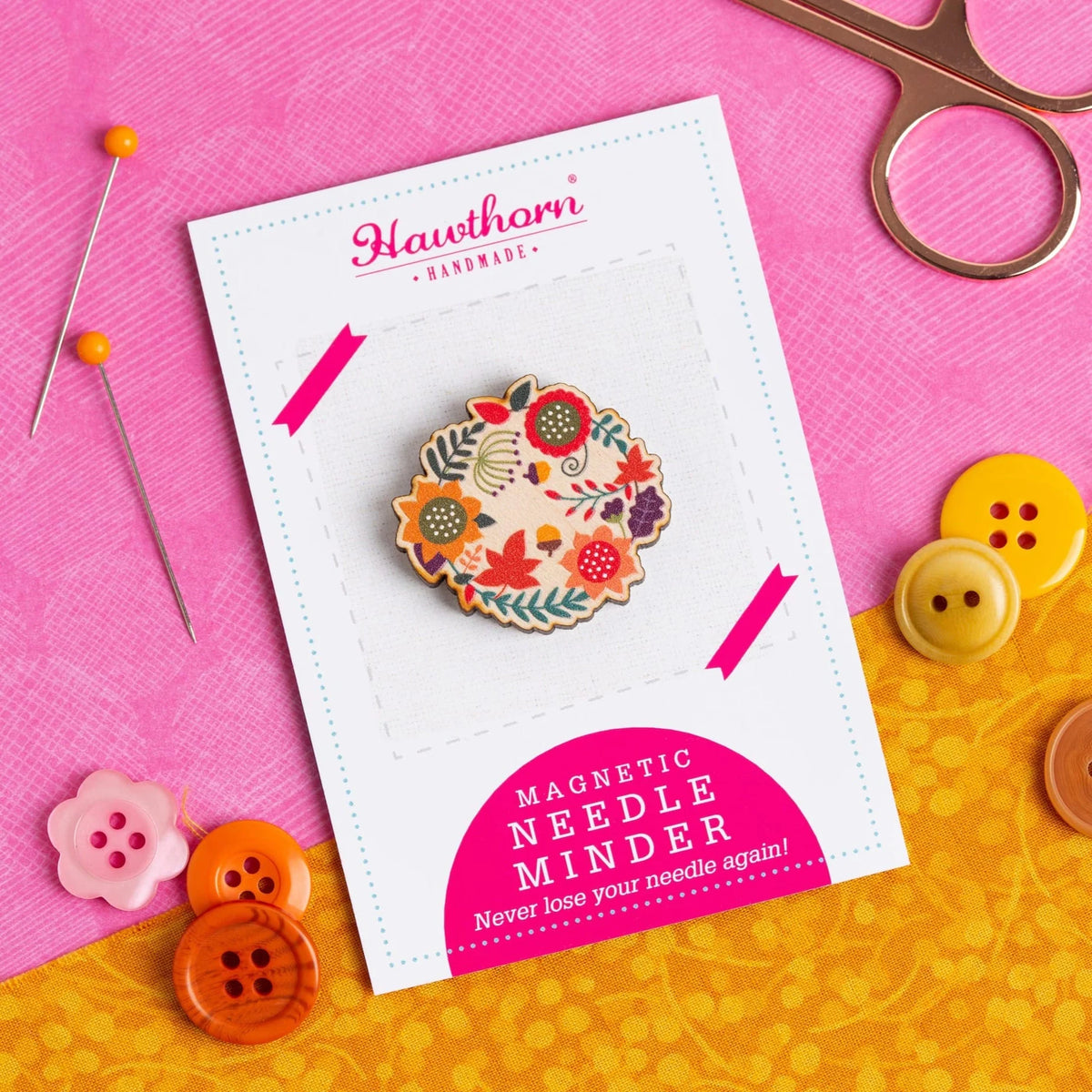 A wooden magnetic needle minder, in an autumn leaves design, on a Hawthorn Handmade label. This placed on pink and yellow fabric, with buttons, pins and scissors surrounding it.