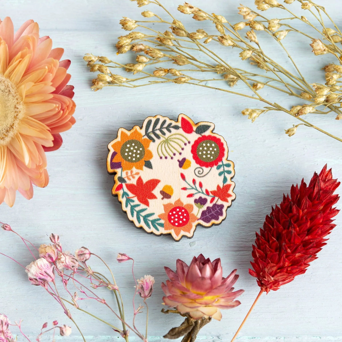 A wooden magnetic needle minder from Hawthorn Handmade, featuring autumn leaves in a colorful hand-painted floral design. Styled in a flat-lay arrangement with dried flowers on a light blue wooden background.