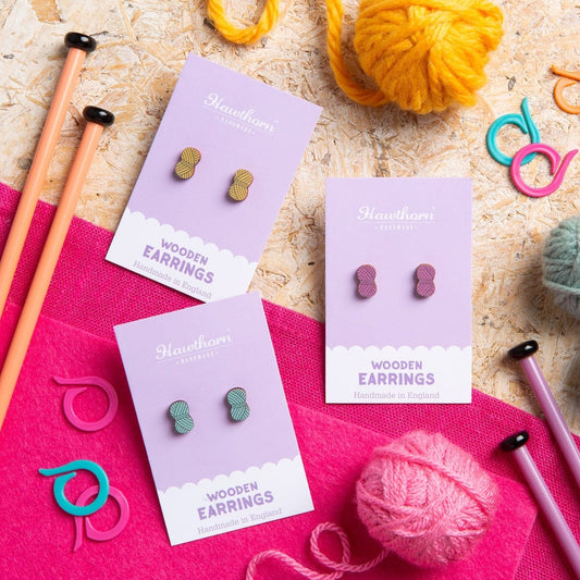 Three pairs of Hawthorn Handmade wooden yarn ball earrings on purple cards with knitting needles and yarn in the background.