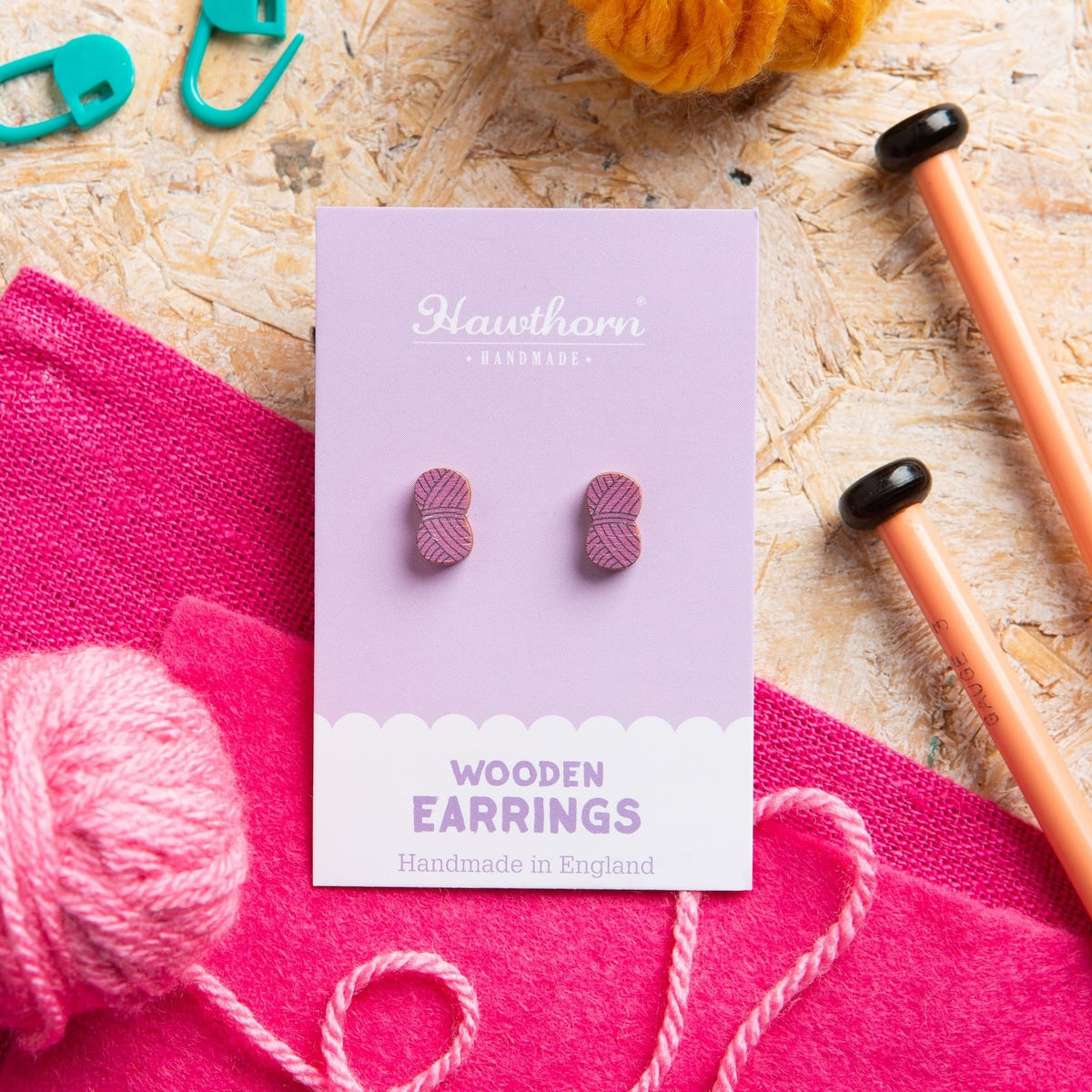 Wooden ball of yarn earrings on a Hawthorn Handmade card with knitting materials in the background.