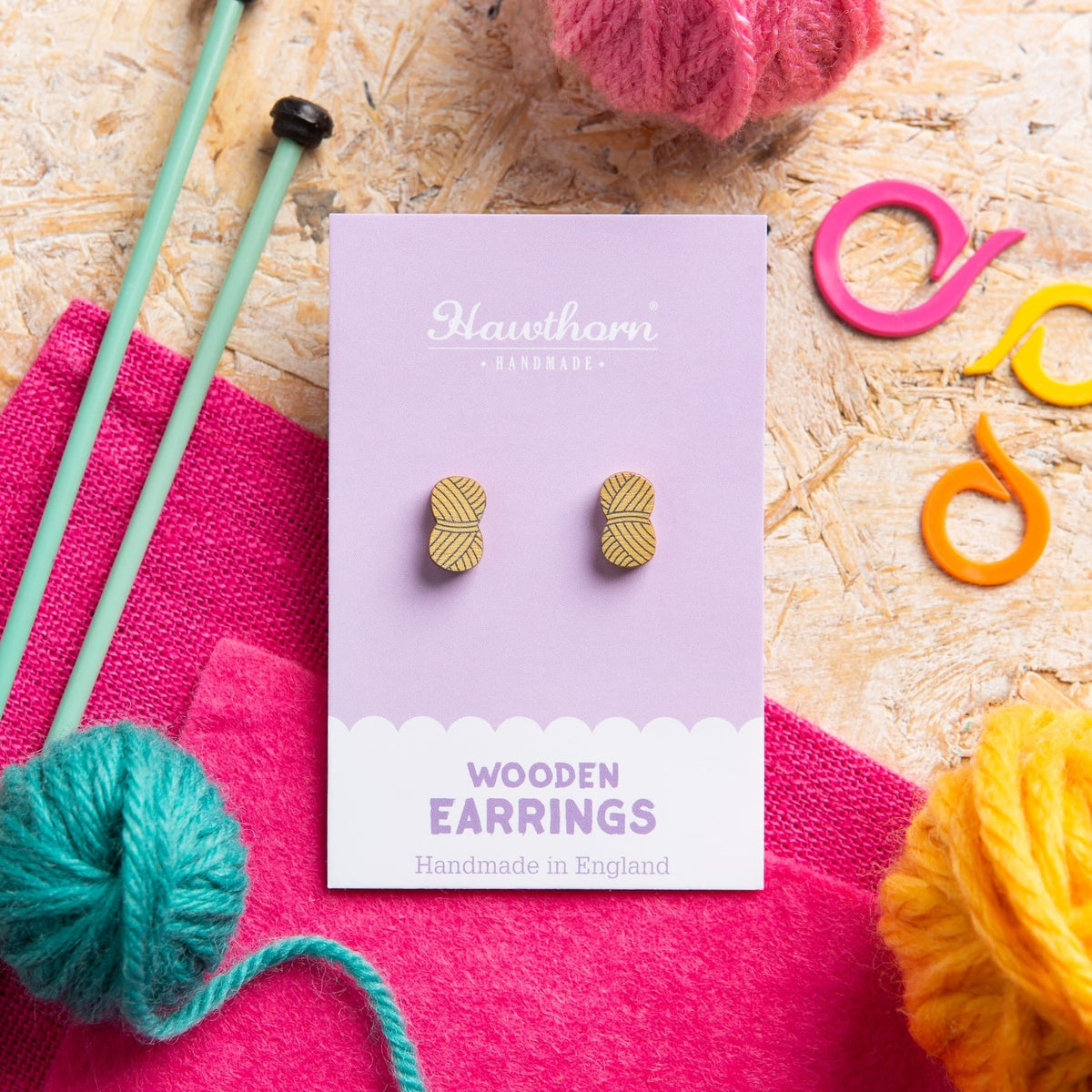 A pair of wooden yarn ball stud earrings in Mustard, displayed on a lavender Hawthorn Handmade card, styled in a flat lay with knitting needles, yarn balls, and colorful crochet hooks on a wood background.