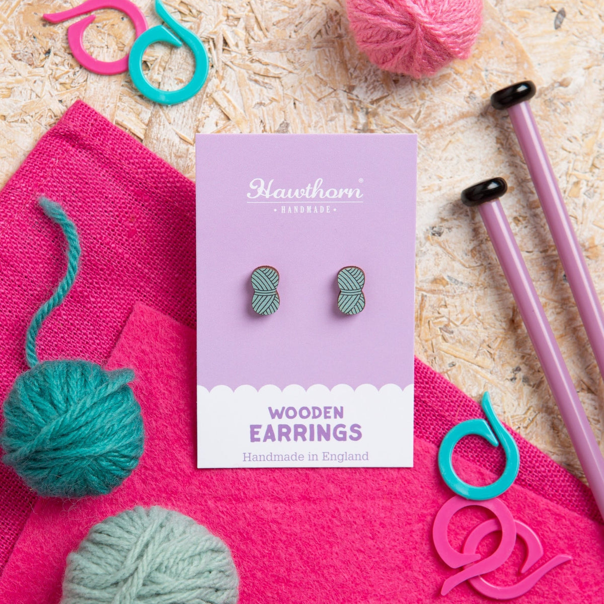 Wooden Teal earrings, shaped like a ball of yarn, on a purple Hawthorn Handmade card, with knitting materials on a textured surface.