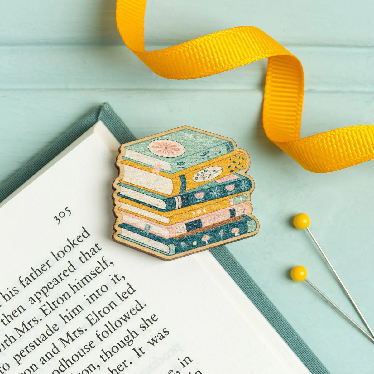 Hawthorn Handmade Book Lover wooden needle minder on an open book with a yellow ribbon and pins.