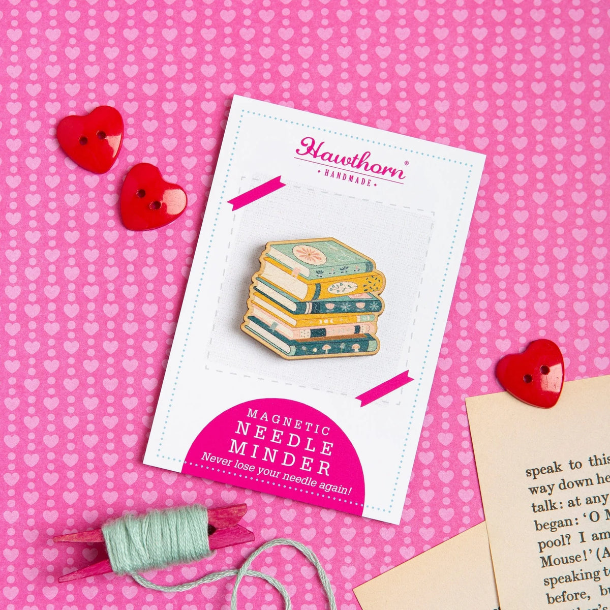 Pile of books wooden magnetic needle minder on Hawthorn Handmade label, placed on pink heart-patterned background.
