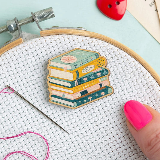 Embroidery hoop with a Book Lover Needle Minder from Hawthorn Handmade on Aida and embroidery needle next to it.