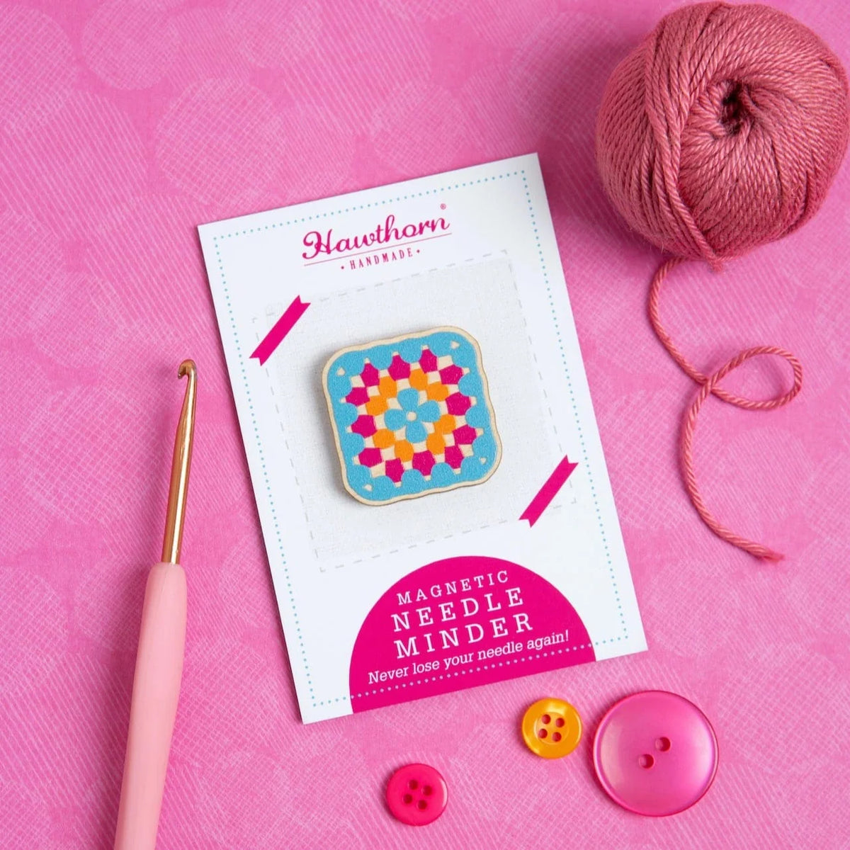 Magnetic needle minder with colourful granny square design, on a Hawthorn Handmade label, next to pink yarn and buttons on a pink background.