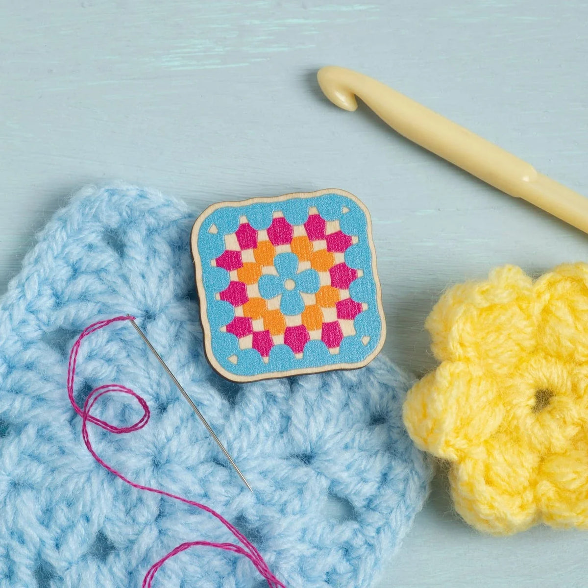 A granny square magnetic needle minder, from Hawthorn Handmade, displayed on a light blue crochet square alongside a yellow crochet hook, a yellow crochet flower, and a needle on a light blue wooden background.