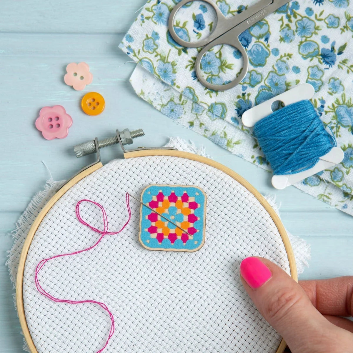 A hand with bright pink nail polish holds a wooden embroidery hoop containing white Aida fabric and needle with pink thread. The needle is magnetised to a wooden needle minder from Hawthorn Handmade, designed as a granny square.