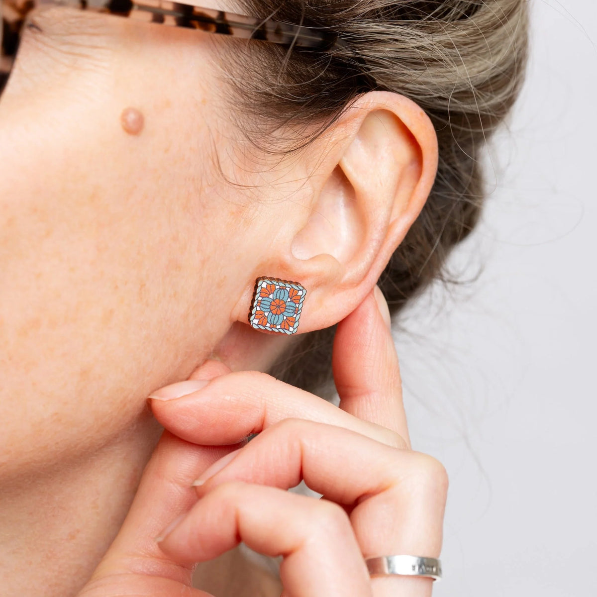 A close-up of a woman's ear wearing a granny square stud earring in turquoise blue, coral, and white.