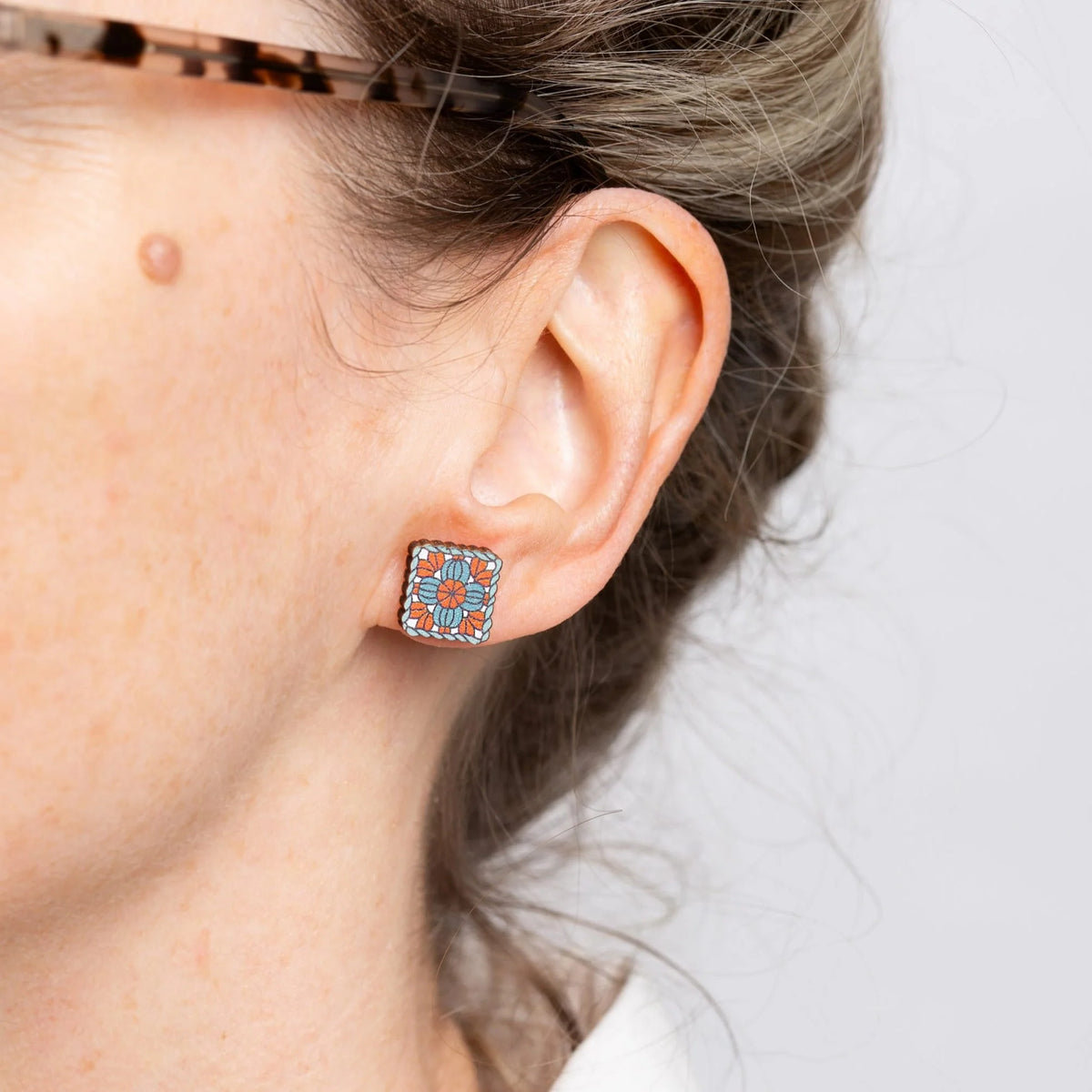 Close-up of an ear wearing a granny square stud earring on a plain background.