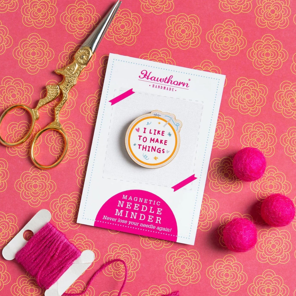 A magnetic needle minder, shaped like an embroidery hoop, with the text 'I Like to Make Things'. This is displayed on a Hawthorn Handmade card, with pink and gold fabric in the background, and gold scissors, pink felt balls, and pink thread next to it.