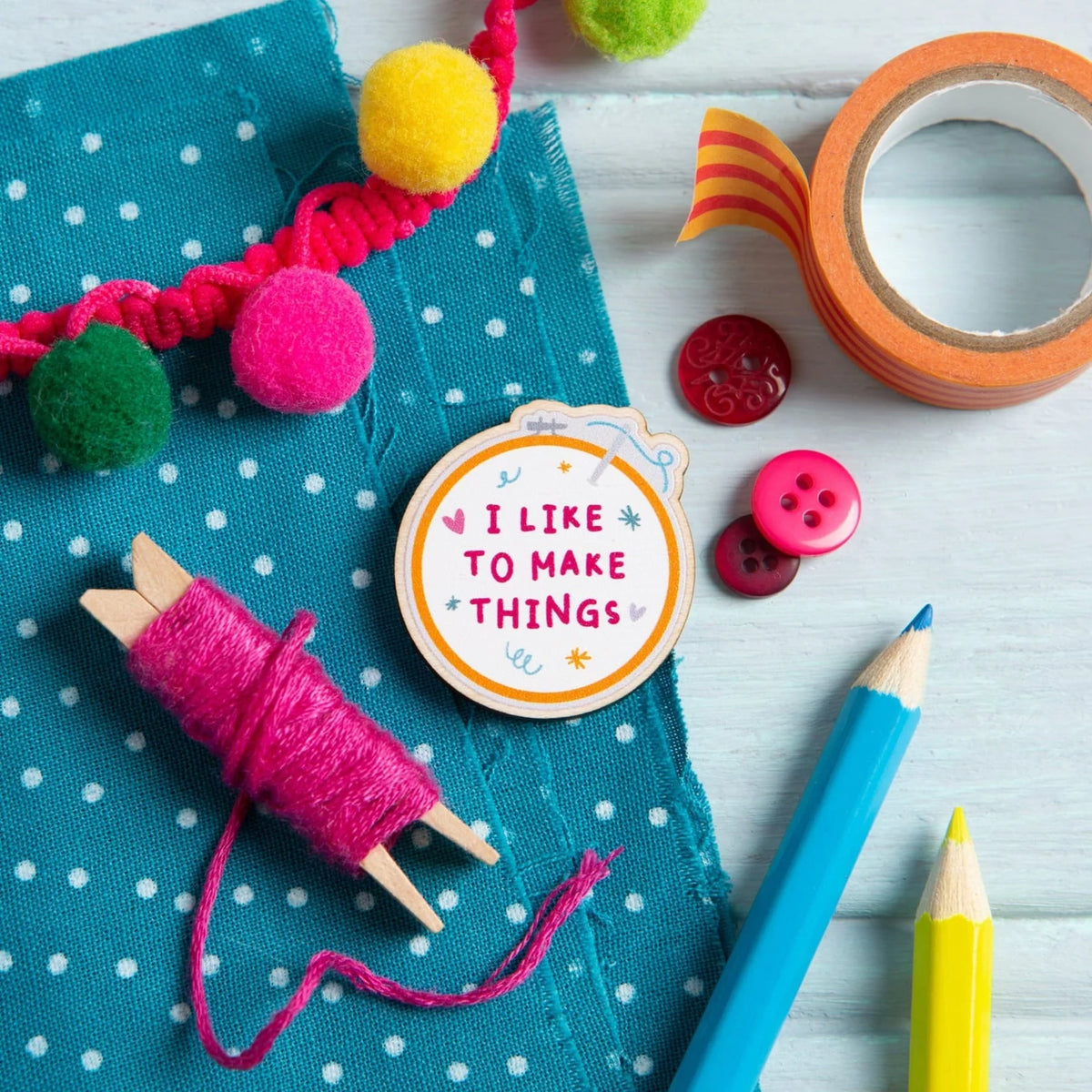 A Hawthorn Handmade magnetic needle minder with the text 'I LIKE TO MAKE THINGS' in pink lettering, surrounded by craft-themed props including a teal polka-dot fabric, pink thread, pom-poms, buttons, washi tape, and colored pencils, on a pale blue wood background.