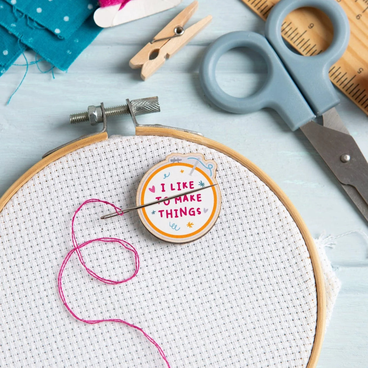 A circular embroidery hoop with white cross-stitch fabric, a magnetic needle minder holding pink thread and a needle holding an embroidery needle. The Hawthorn Handmade wooden needle minder is shaped like an embroidery hoop and reads “I LIKE TO MAKE THINGS.” Scissors, a wooden clothespin, fabric, and a ruler are arranged on a pale blue surface.