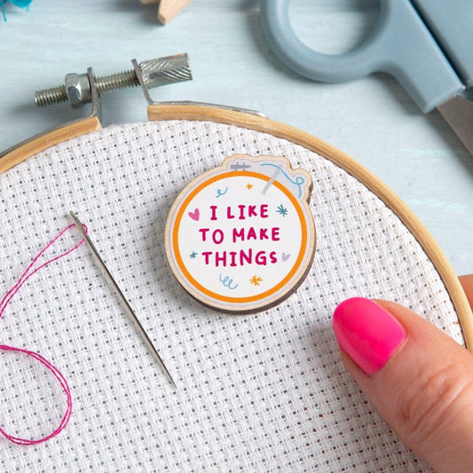 An embroidery hood themed needle minder, with the text 'I Like to Make Things'. The needle minder is placed on an embroidery hoop with Aida fabric, with an embroidery needle and pink thread next to it.