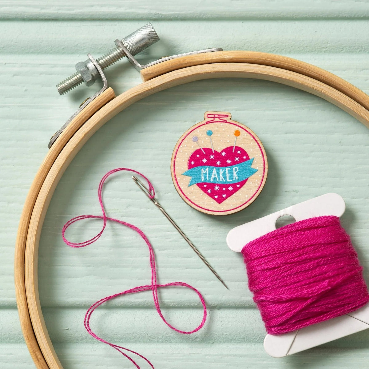 A wooden embroidery hoop arranged on a light mint green wooden surface, alongside pink thread, an embroidery needle, and a Hawthorn Handmade needle minder featuring a pink heart and the word "MAKER" on a blue banner.
