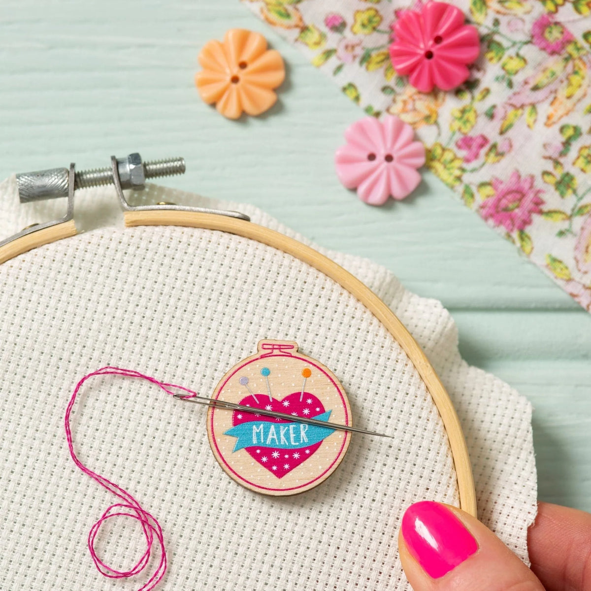 A close-up of a hand with bright pink nails holding a wooden embroidery hoop with Aida fabric. A needle threaded with pink thread is magnetised to a needle minder that reads “MAKER” inside a pink heart pin cushion.