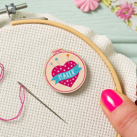 A close-up of a circular wooden embroidery hoop holding cream fabric, with a hand holding the hoop. An embroidery needle with pink thread rests next to a needle minder from Hawthorn Handmade. The design is a pink heart pin cushion with white dots and a blue banner reading “MAKER".