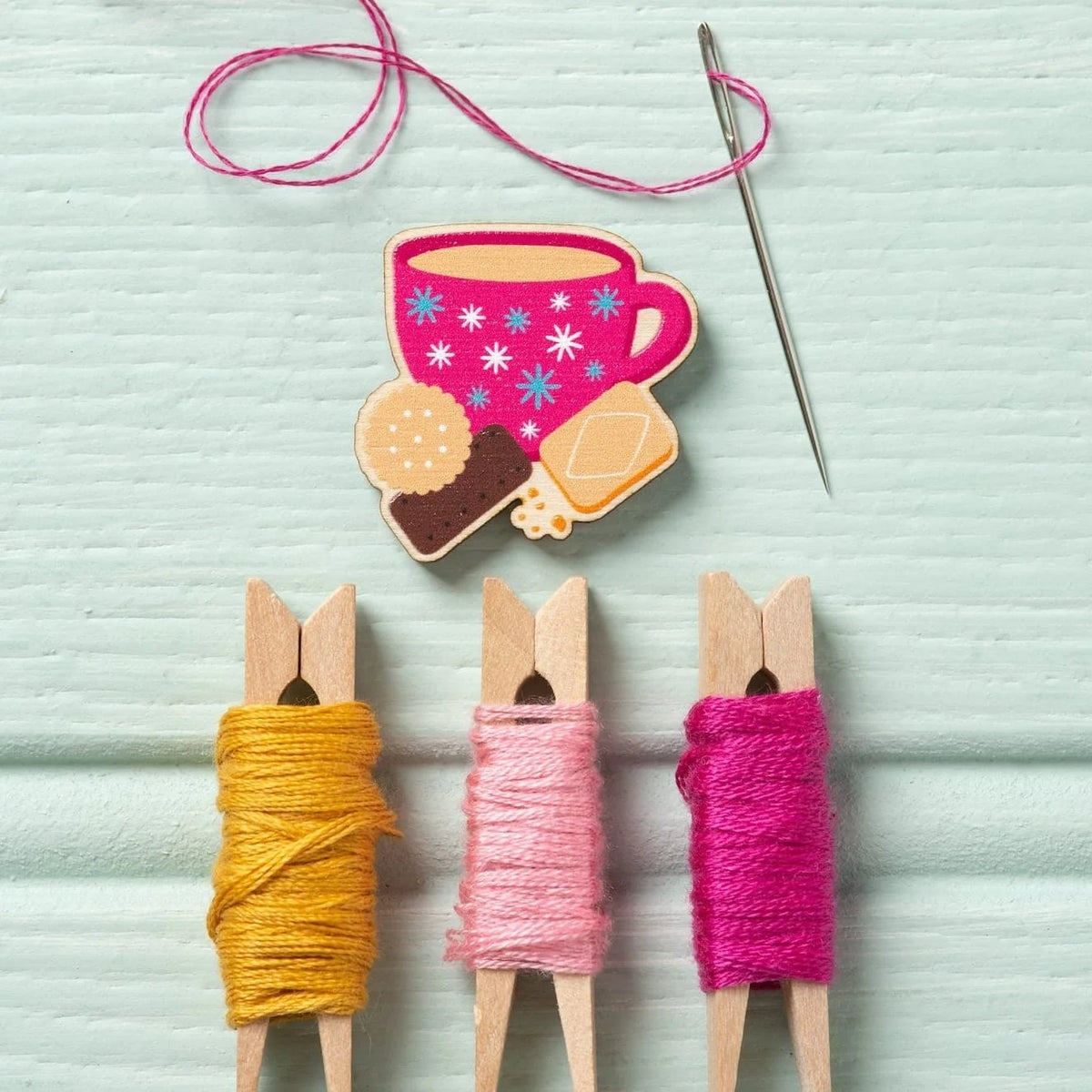 A blue wooden background, with an embroidery needle threaded with pink thread. Below is a magnetic needle minder from Hawthorn Handmade, shaped like a pink teacup and biscuits. Below this are 3 clothes pegs wrapped with pink and yellow embroidery threads.