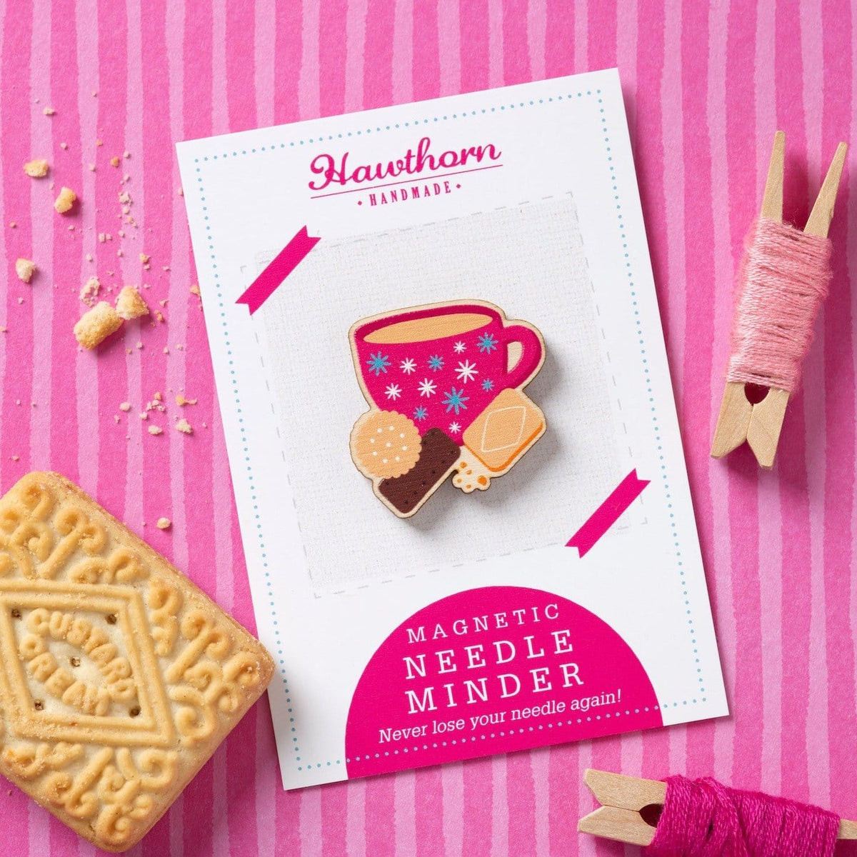 A wooden magnetic needle holder, shaped like a cup of tea and biscuits, on a Hawthorn Handmade card. The background is pink striped fabric, with pegs wrapped in embroidery thread and a biscuit to the side.