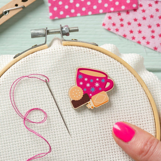 A close-up of a wooden embroidery hoop holding Aida fabric, with a colorful magnetic needle minder shaped like a pink tea cup and biscuit. A hand with bright pink nail polish is visible at the lower right, and embroidery thread and a needle appear on the left.