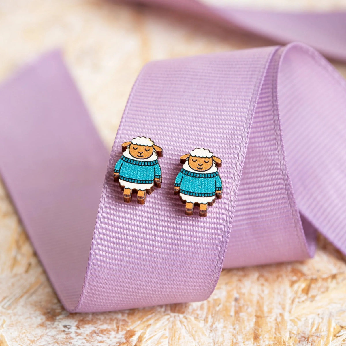 Woolly Sheep wearing sweaters earrings on a purple ribbon with a natural background.