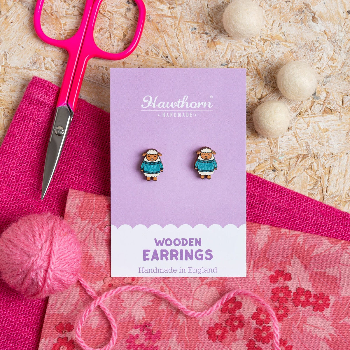 Wooden Woolly Sheep earrings on a Hawthorn Handmade card with pink fabric, pink yarn and scissors on a textured surface.