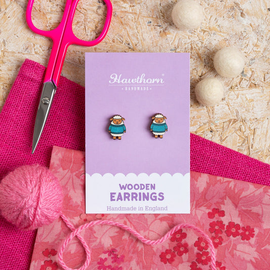 Wooden Woolly Sheep earrings on a Hawthorn Handmade card with pink fabric, pink yarn and scissors on a textured surface.