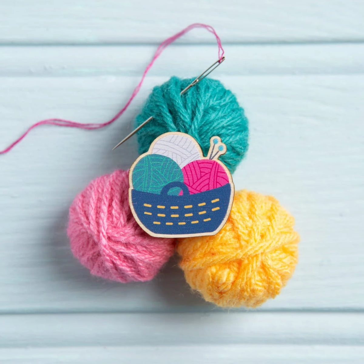 Magnetic needle minder, shaped like a basket of yarn, from Hawthorn Handmade. The needle minder is placed on 3 small balls of yarn, in pink, turquoise and yellow. There is an embroidery needle sticking out of the turquoise yarn ball. The background is pale blue wood.
