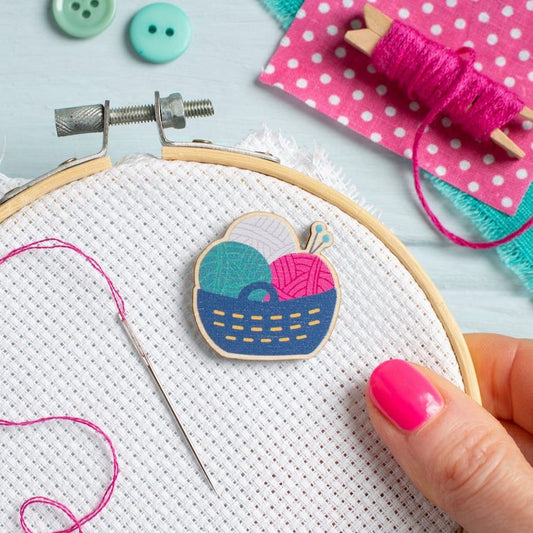 Hawthorn Handmade needle minder, shaped like a basket of yarn. Placed on a piece of Aida in an embroidery hoop, with an embroidery needle next to it, and craft supplies to the side.