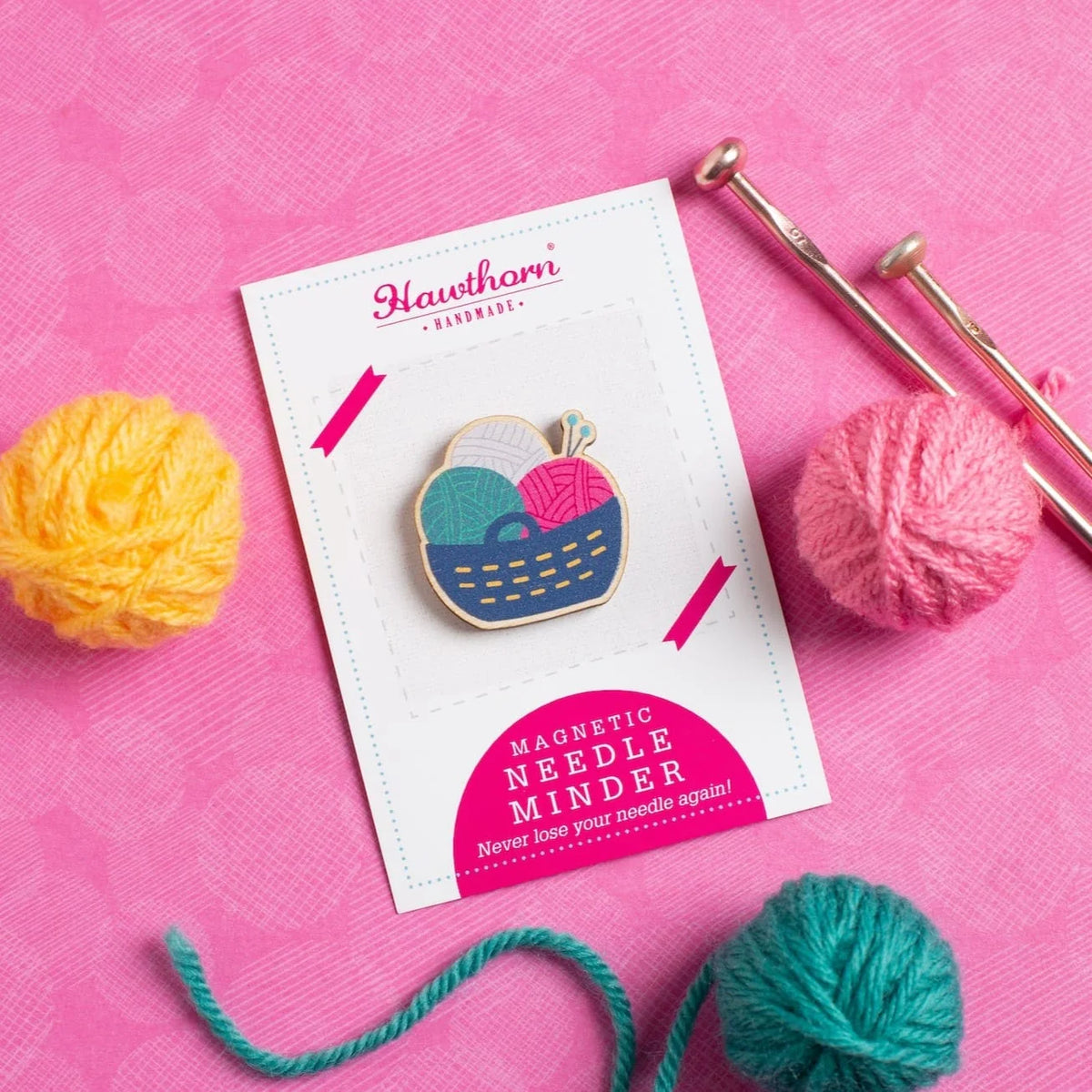 Wooden yarn basket needle minder, set on a Hawthorn Handmade label, with balls of wool and knitting needles to the sides. The background is pink fabric.