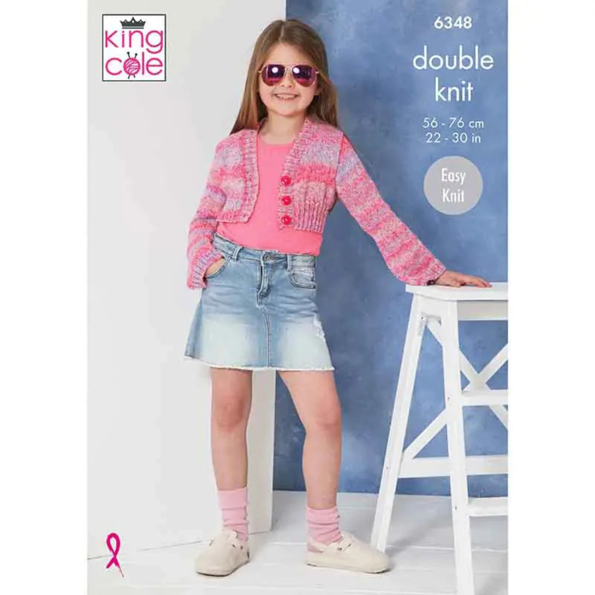 King Cole easy knit dk pattern featuring a young girl wearing a pink knitted cardigan and denim skirt, standing next to a white stool.The pattern is 6348.