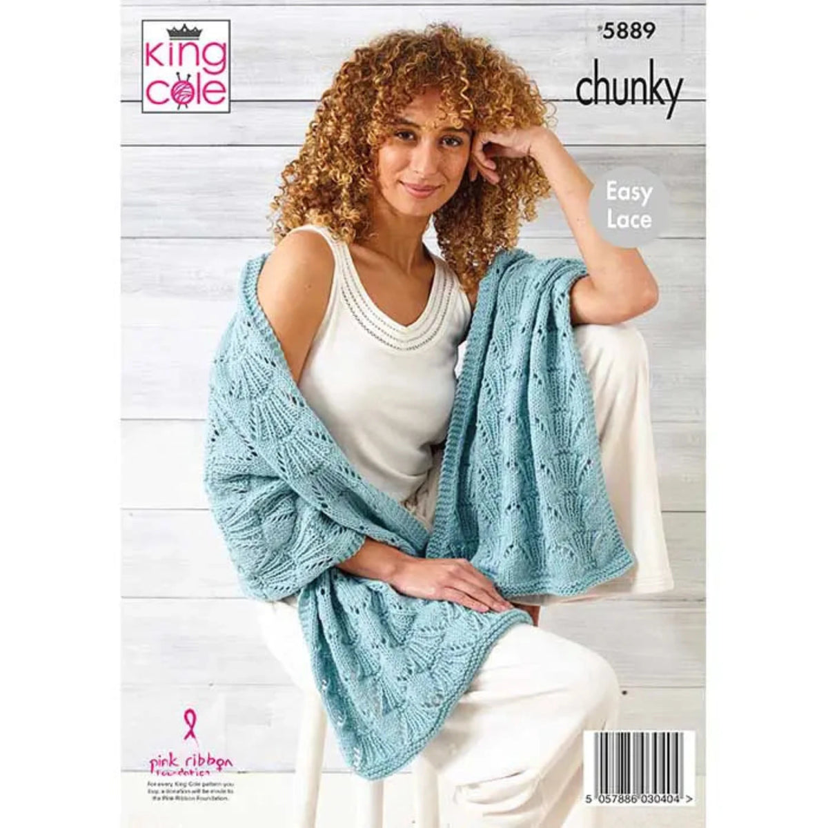 A woman seated against a light wood plank wall wearing a chunky light blue knitted, easy lace wrap, over a white sleeveless top and white pants.