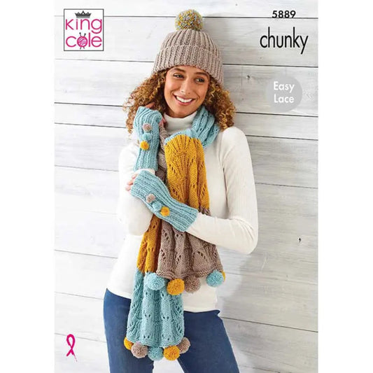 A woman wearing a beige knit beanie with a pom-pom, a white sweater, and blue jeans models a chunky knit scarf and matching glove set. She is stood against a light wood plank background. The scarf and gloves feature a mix of light blue, mustard yellow, taupe brown, and cream colors with pom-pom accents.