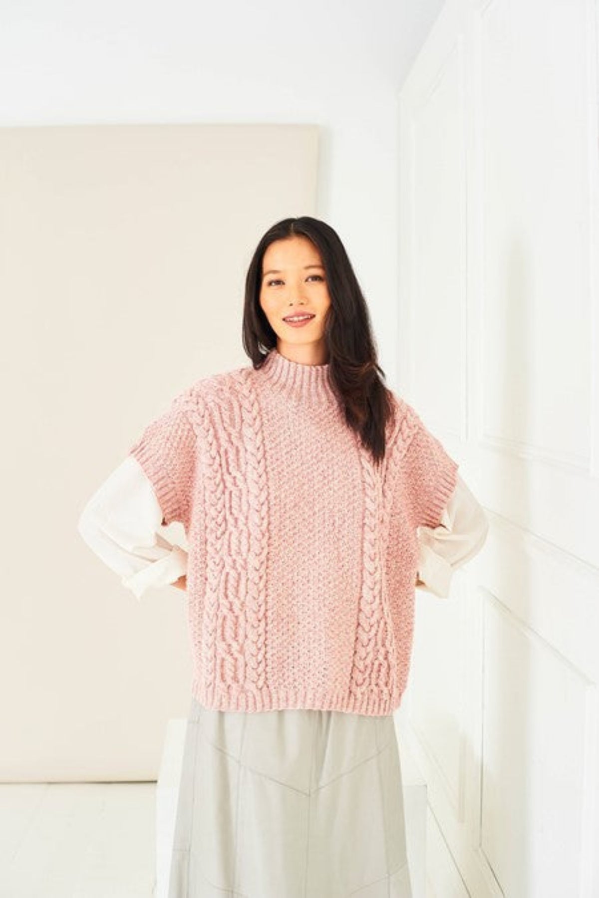 A woman wearing a textured cable tank top sweater, knitted in Stylecraft Life DK yarn from the pattern 10284. The top is in shade Rose Wash. The model is also wearing a white long sleeve tee shirt and beige skirt.