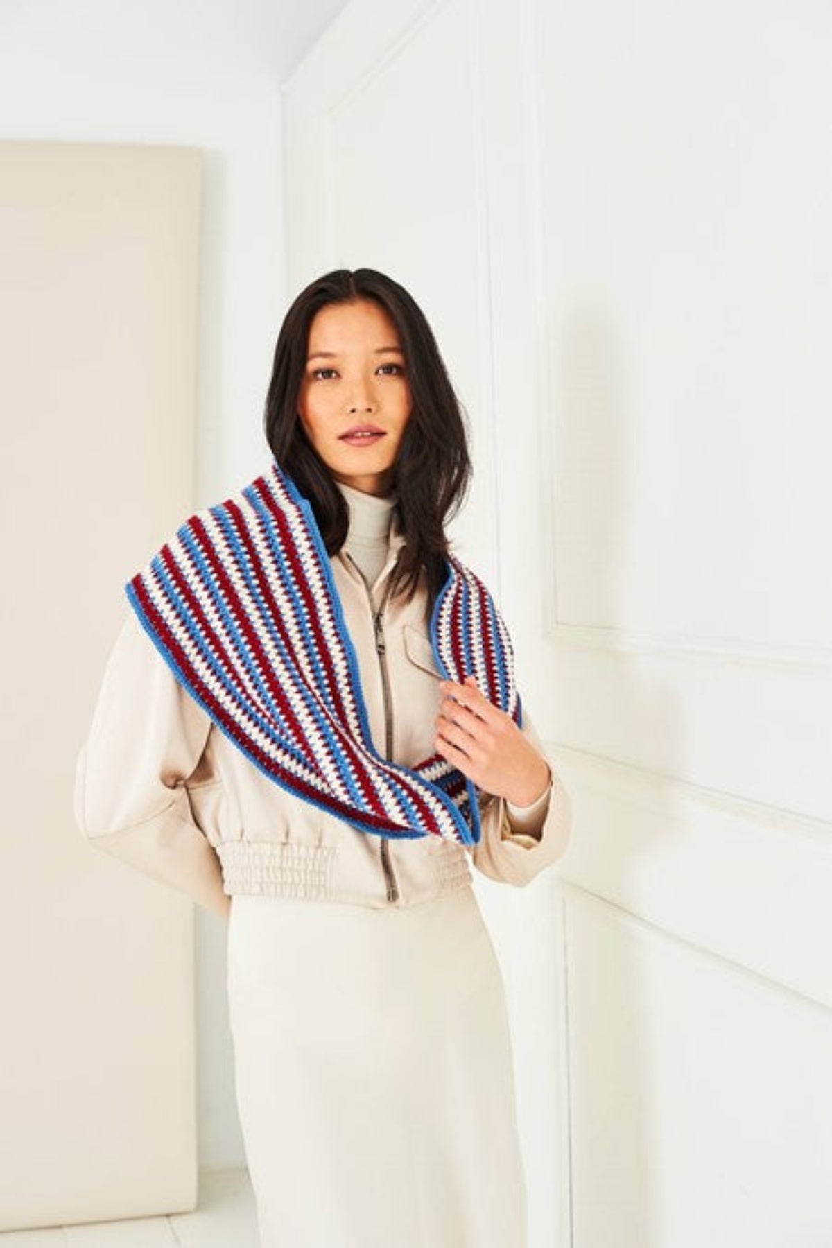 A woman wearing a cream-colored jacket and white skirt stands in a bright white room, draped with a crochet cowl  scarf featuring red, blue, and white stripes. The cowl is rom Stylecraft pattern 10287, and is crocheted Naturals Pure Wool DK yarn, shades Cream, Claret and Denim.