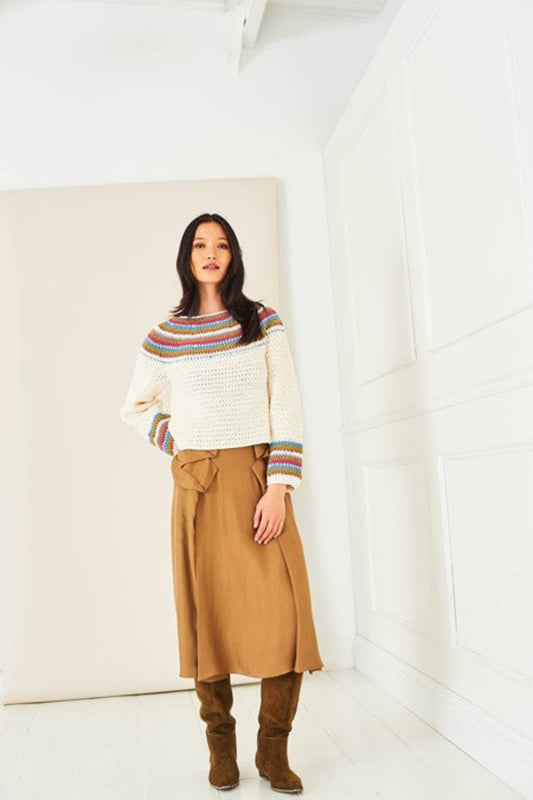 A women modelling a yoke crochet sweater from Stylecraft pattern 10287. She is stood in a bright white room and the sweater features stripes at the neckline and the cuffs. The sweater is crocheted in Stylecraft Naturals Pure Wool DK yarn, in shades Raspberry, Cream, Mistletoe, Storm Blue and Mocha. She is also wearing a mid length tan skirt and brown suede boots.