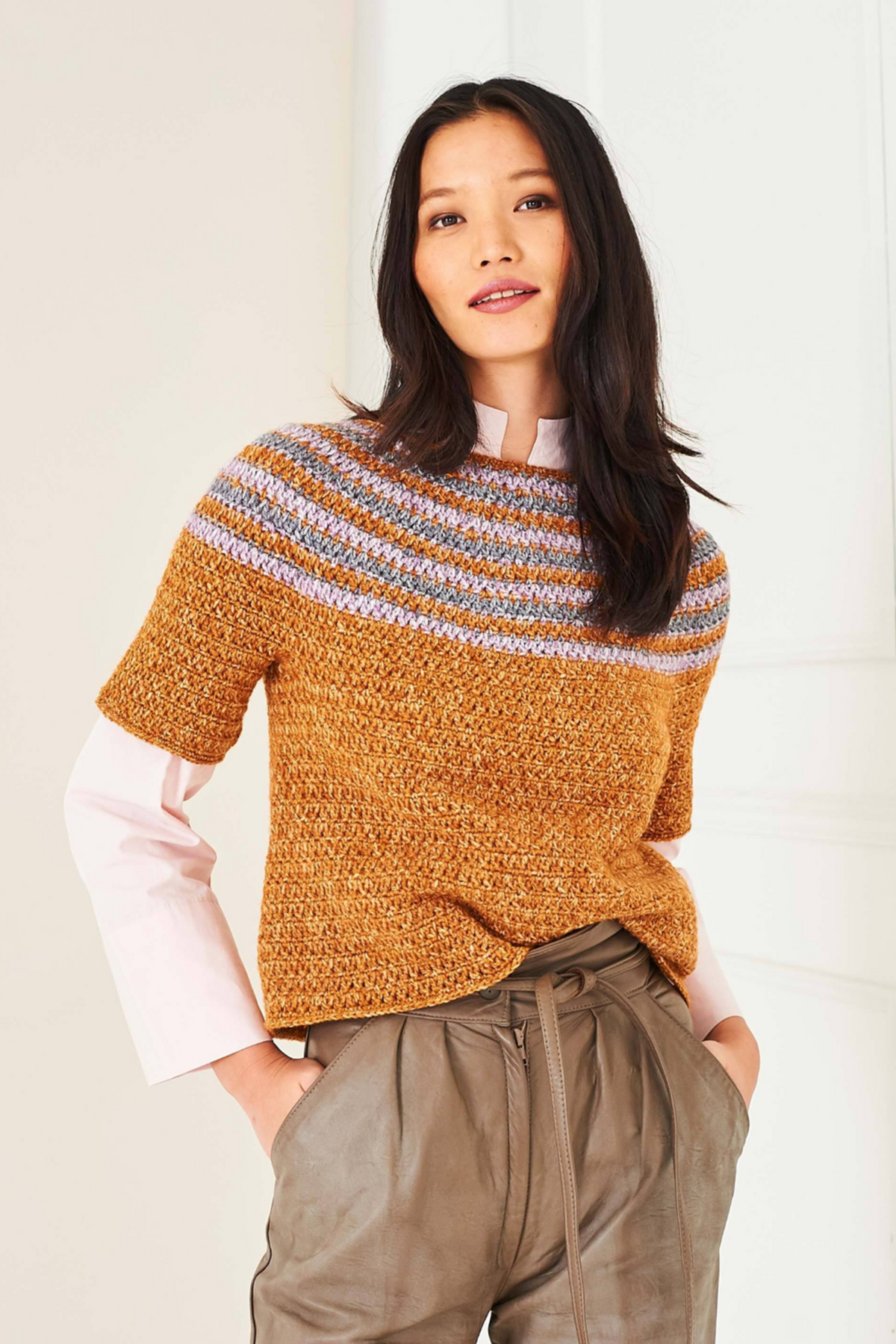 A women against a white background is modelling a yoke crochet top from Stylecraft pattern 10287. The top features stripes at the neckline. The top is crocheted in Stylecraft Life DK yarn, in shades Ginger Wash, Graphite Wash and Heather Wash. She is also wearing a pale pink shirt and light brown faux leather trousers.