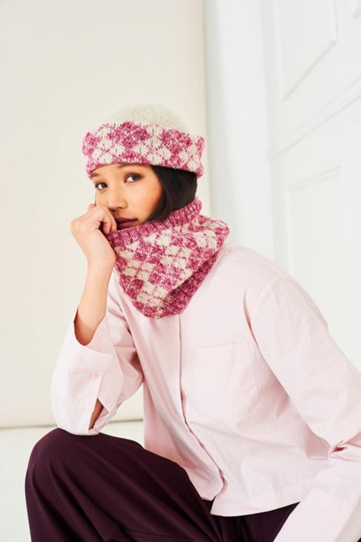 A woman wearing a pink and cream Faire Isle knitted hat and matching neck warmer cowl, paired with a light pink shirt and dark purple pants, sitting in a brightly lit room. The hat and cowl are knit in Stylecraft Life DK, shades Raspberry Wash and Parchment, and the pattern is 10285.