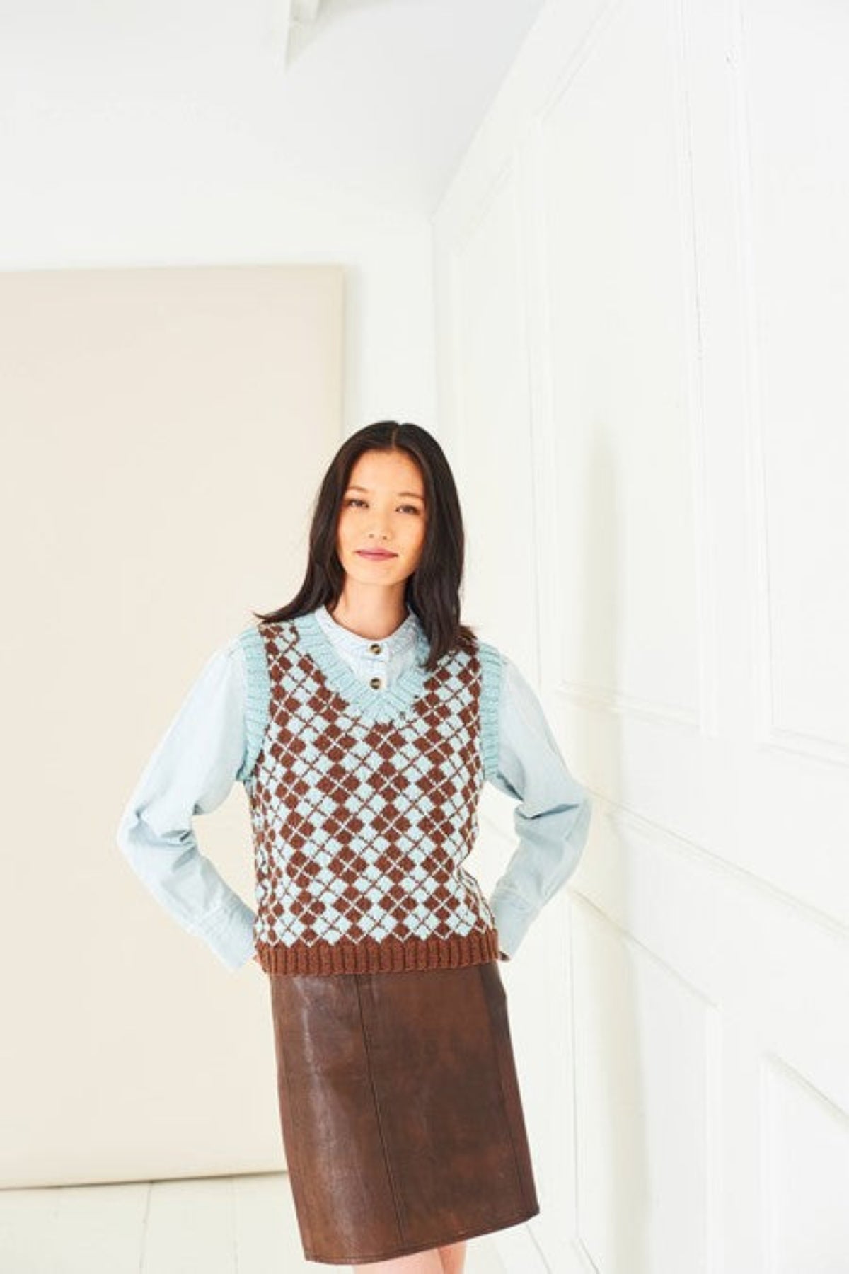A woman wearing a Faire Isle tank top knitted in Stylecraft Naturals Pure Wool DK yarn, shades Walnut and Duck Egg. She is also wearing a brown faux leather skirt and a pale blue shirt. The tank top is from Stylecraft pattern 10285