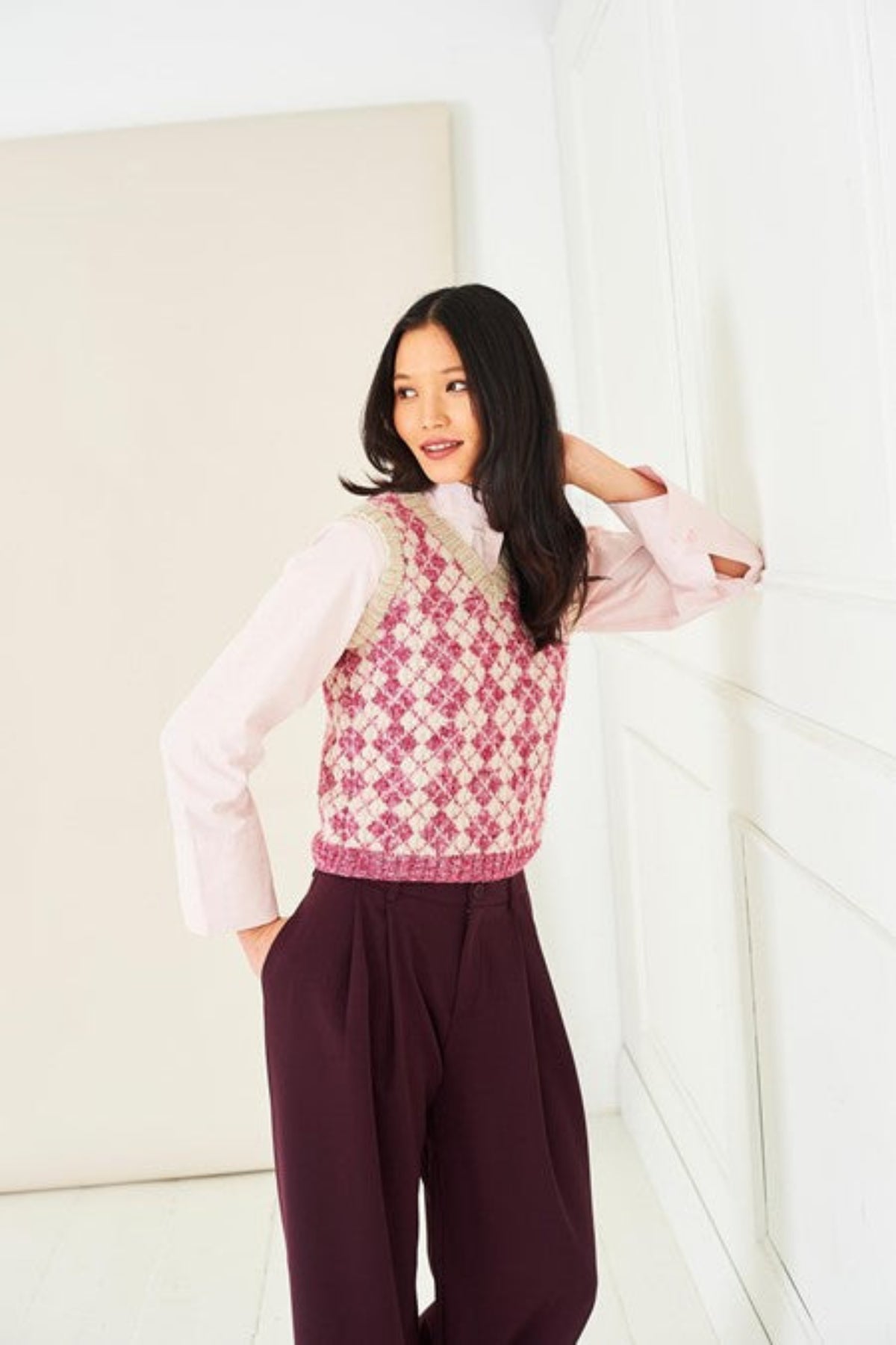 A woman wearing a Faire Isle tank top knitted in Stylecraft Life DK yarn, shades Raspberry Wash and Parchment. She is also wearing purple trousers and a pale pink shirt. The top is from Stylecraft pattern 10285.