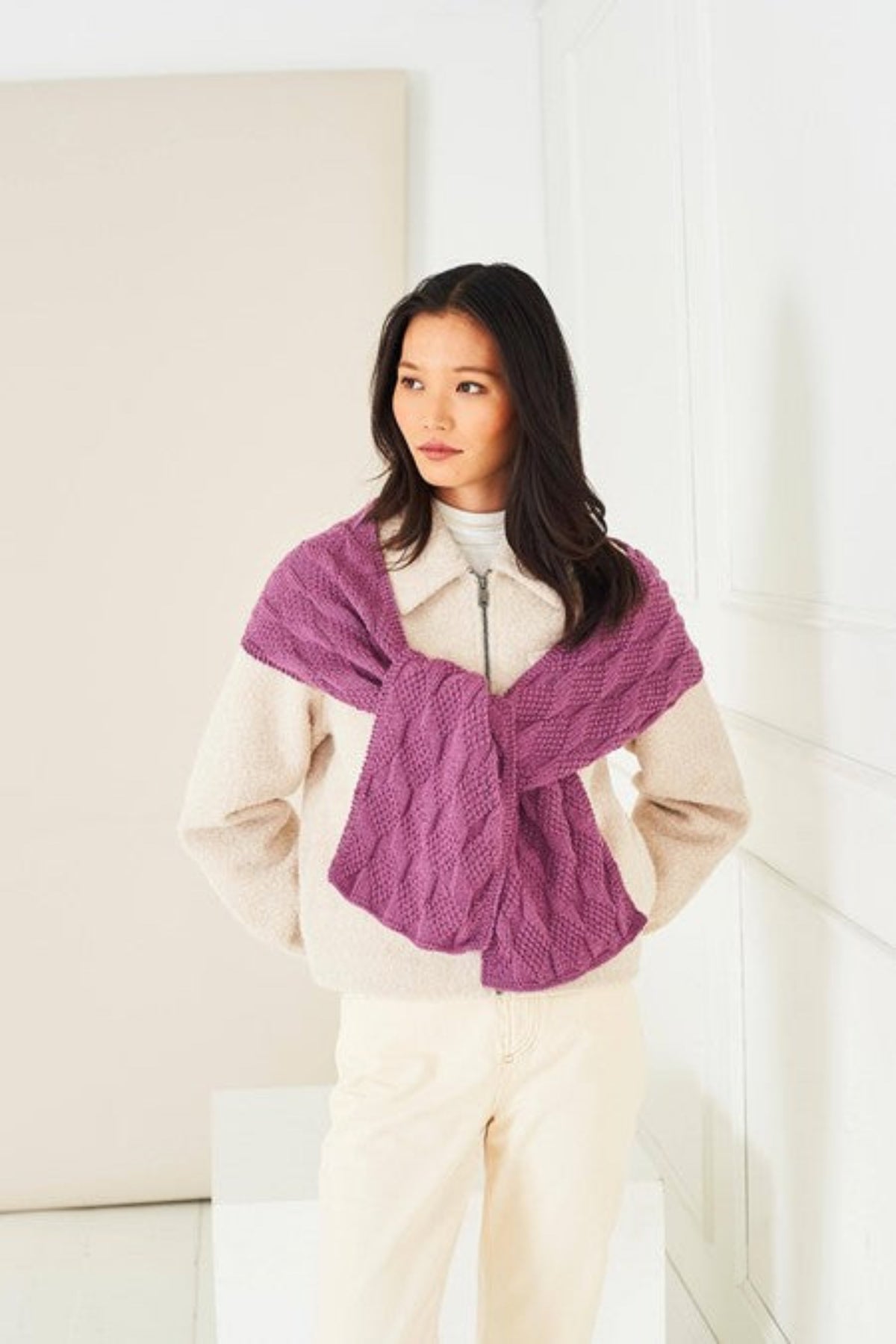 A woman wearing a cream-colored zip-up jacket and cream pants, wrapped in a textured knit purple scarf, knitted in Stylecraft Naturals Pure Wool DK yarn in shade Grape. She is standing in a bright, minimalist room with white panelled walls.