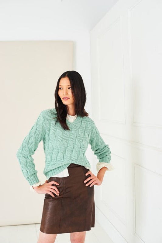 A woman modelling a mint green textured sweater, knit in Stylecraft Naturals Pure Wool DK yarn in shade Lincoln. The sweater is worn over white collared shirt and a brown faux-leather mini skirt, standing in a bright white room with panelled walls.