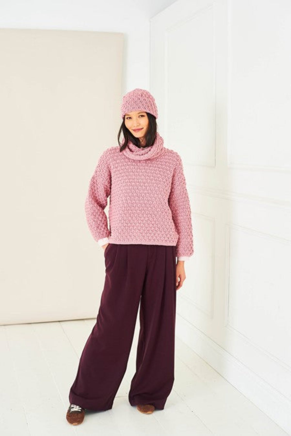A woman stands in a bright white room wearing a pink crochet cowl-neck textured sweater, and a matching pink crochet beanie. She also is wearing dark purple wide-leg trousers. The hat and sweater are from Stylecraft pattern 10286.