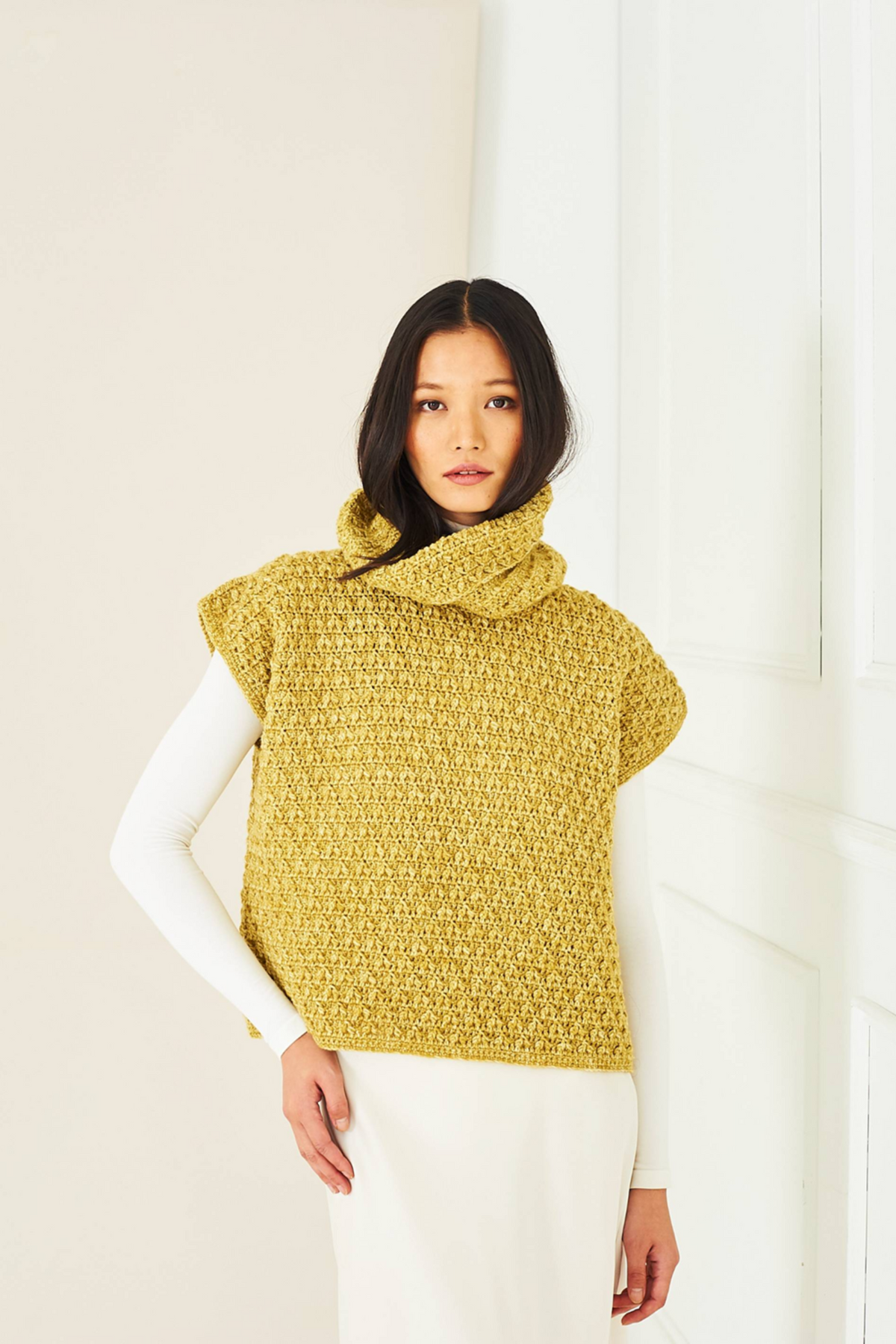 A woman stands in a bright white room wearing a yellow crochet cowl-neck textured tank top. She also is wearing a long sleeve tight white dress. The tank top is from Stylecraft pattern 10286.