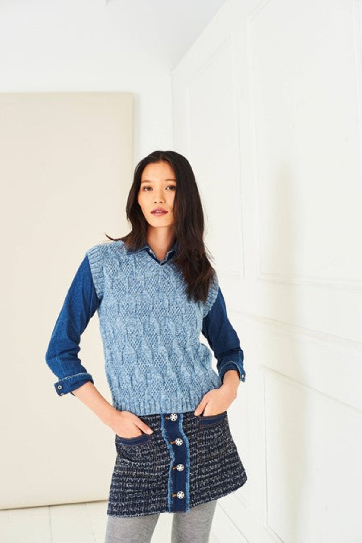 A woman wearing a v-neck knitted tank top rom Stylecraft pattern 10282. The tank top is knitted in Stylecraft Life DK yarn in shade Storm Wash, which is a shade of blue. She wears this over a denim shirt and a tweed skirt. The background is a white panelled room.