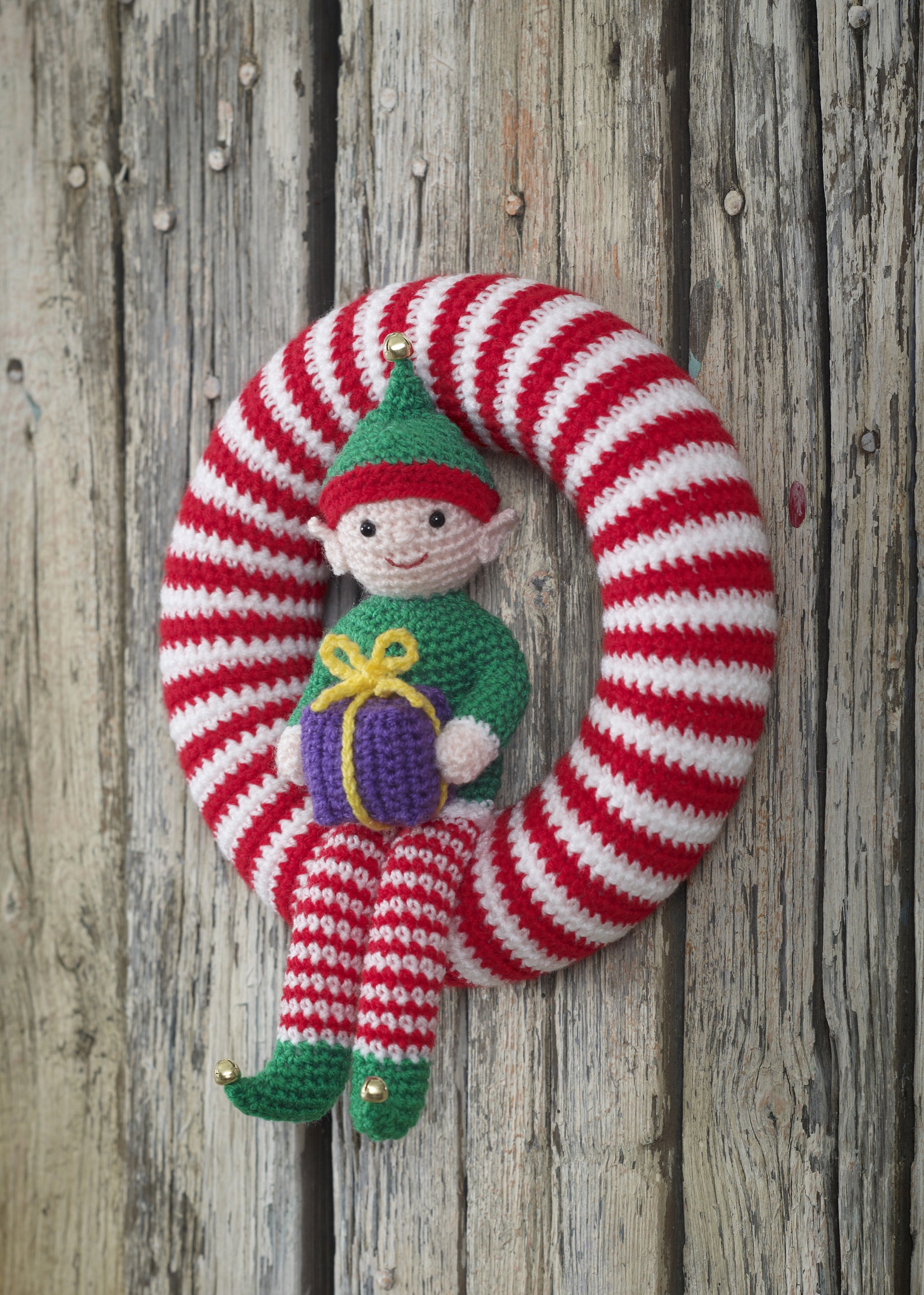 Colourful elf wreath to decorate. A red and white striped wreath with a red and green elf sat on holding a purple gift. This wreath is approximately 78.5cm around the outside of the ring to fit a polystyrene ring of this size.